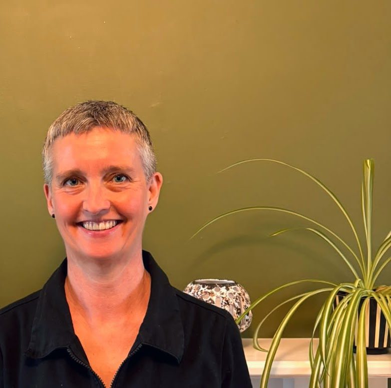 Vic, smiling, in front of a green wall with a spider plant
